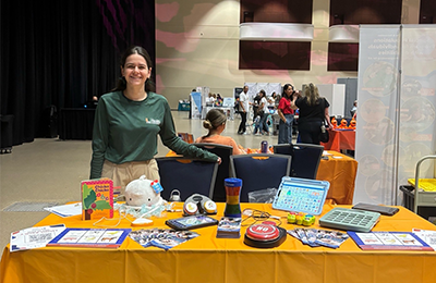 A white woman with long brown hair smiles, standing behind an exhibit booth full of materials.